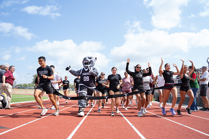 General the Jaguar and Student-Athletes Running Through Finish Line Ribbon
